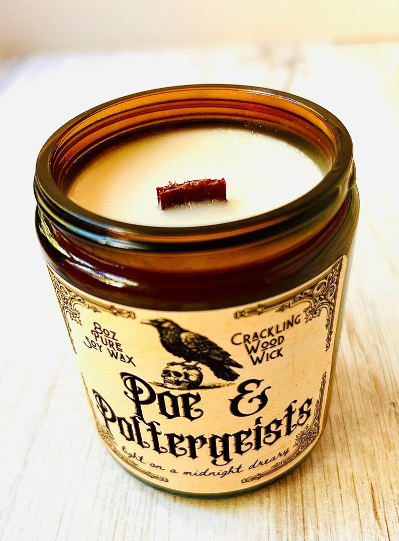 K&ouml;nnte beinhalten: Braune Glas-Kerze mit Holzdocht. Das Etikett zeigt einen schwarzen Raben &uuml;ber einem Totenkopf mit den Worten "Poe & Poltergeists". Die Kerze enth&auml;lt 227g reines Sojawachs und einen knisternden Holzdocht.