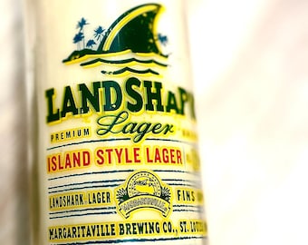 May include: A clear glass bottle filled with a creamy liquid, labeled "LANDSHARK Lager." The label features a shark fin graphic and text that reads "Island Style Lager" and "Margaritaville Brewing Co., St. Louis."