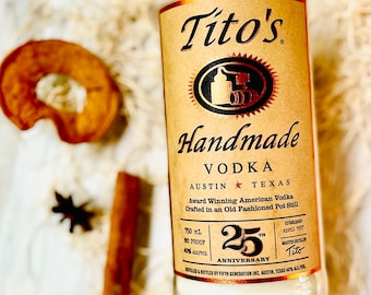 May include: A bottle of Tito's Handmade Vodka, with a tan label and gold accents, is the focal point. The label includes text, a logo, and details about the product. A dried apple slice, a cinnamon stick, and a star anise are also visible.