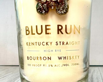 May include: A clear glass candle with a cream-colored wax. The candle has gold lettering that reads "BLUE RUN" and "KENTUCKY STRAIGHT BOURBON WHISKEY". A silver butterfly adorns the top. The candle is 750ml.