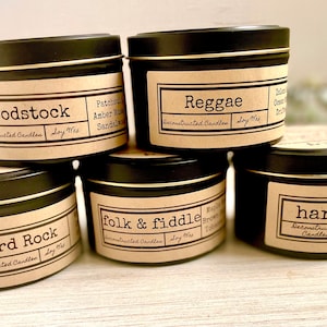 May include: Six black candle tins with gold trim, stacked in a pyramid shape. Each tin has a brown paper label with a different name and scent description. The names are "Woodstock", "Reggae", "Hard Rock", "Folk & Fiddle", "Harmony", and "Mountain Brew".