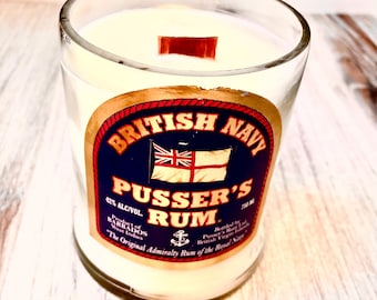 May include: A clear glass candle jar with a white candle and a wooden wick. The jar has a label that says "British Navy Pusser's Rum" with a red, white, and blue flag. The label also says "42% Alc/Vol" and "Product of Barbados West Indies".