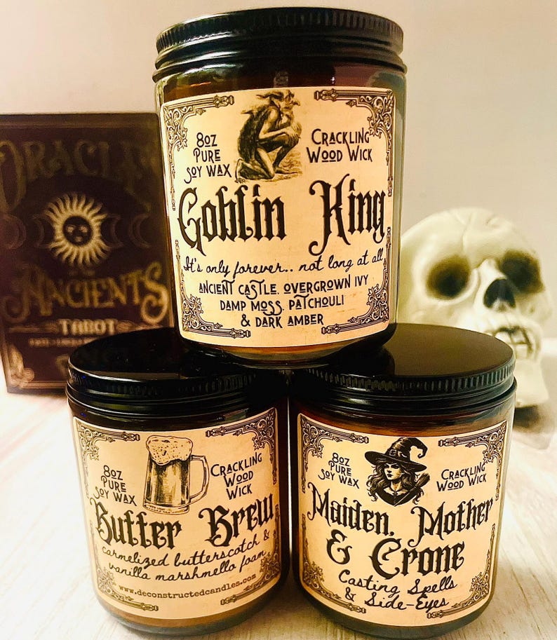 K&ouml;nnte beinhalten: Drei Sojawachskerzen in Gl&auml;sern mit schwarzen Deckeln. Die Etiketten zeigen Illustrationen und Text, darunter "Goblin King", "Butter Brew" und "Maiden, Mother & Crone". Jede Kerze ist 227g und hat einen knisternden Holzdocht.