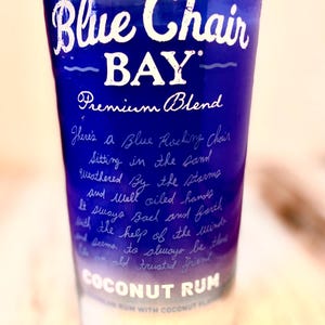 May include: A blue glass bottle with a white label that reads "Blue Chair BAY Premium Blend Coconut Rum". The label also has a handwritten message about a blue rocking chair sitting in the sand.