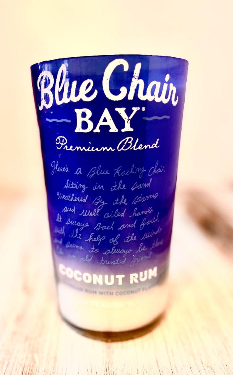 May include: A blue glass bottle with a white label that reads "Blue Chair BAY Premium Blend Coconut Rum". The label also has a handwritten message about a blue rocking chair sitting in the sand.