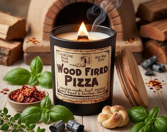 Puede incluir: Una vela negra encendida con una mecha de madera, etiquetada como "Wood Fired Pizza". La etiqueta enumera ingredientes como humo de fogata, albahaca y orégano. La vela está rodeada de hojas de albahaca, un pequeño panecillo y un horno de pizza en miniatura.