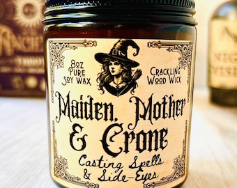 May include: A glass jar candle with a black lid and a label featuring a witch illustration. The label reads "Maiden, Mother & Crone" and "Casting Spells & Side-Eyes." The candle contains 8 oz of pure soy wax and has a crackling wood wick.