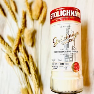 以下が含まれることがあります： 白いラベルと赤いアクセントが付いた透明なガラス瓶のストリチナヤ・ウォッカ。ラベルには「The Original Stolichnaya Vodka」と「Made with Certified Alpha Grade Spirit」と書かれている。瓶は750mlで、ロット番号は12712。瓶は木製の表面の上に置かれている。