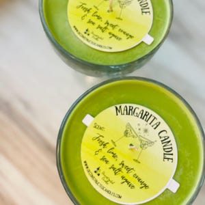 May include: Two green glass margarita candle jars with white labels. The labels have a yellow watercolour background and say "MARGARITA CANDLE" in black script. The labels also say "SCENT: Fresh lime, sweet orange & sea salt agave" in black script.