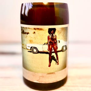 May include: A brown glass bottle with a vintage photograph of a woman standing next to a white car in a desert setting. The woman is wearing a swimsuit and a vest. The photograph is faded and has a yellowed tint.