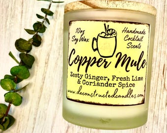 Może przedstawiać: Świeca sojowa o pojemności 10 uncji z drewnianym wieczkiem. Etykieta jest żółta z czarnym tekstem, który brzmi: "Handmade Cocktail Scents Copper Mule Zesty Ginger, Fresh Lime & Coriander Spice www.deconstructedcandles.com".
