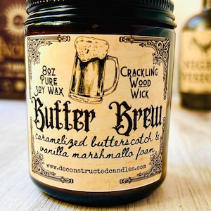 K&ouml;nnte beinhalten: Eine dunkle Glas-Kerze im Glas mit schwarzem Deckel. Das Etikett lautet "Butter Brew" mit "8oz Pure Soy Wax" und "Crackling Wood Wick". Die Kerze zeigt eine Zeichnung eines Bierkrugs und listet den Duft als Karamell-Butterscotch und Vanille-Marshmallow-Schaum auf.