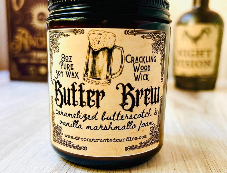 K&ouml;nnte beinhalten: Eine dunkle Glas-Kerze im Glas mit schwarzem Deckel. Das Etikett lautet "Butter Brew" mit "8oz Pure Soy Wax" und "Crackling Wood Wick". Die Kerze zeigt eine Zeichnung eines Bierkrugs und listet den Duft als Karamell-Butterscotch und Vanille-Marshmallow-Schaum auf.