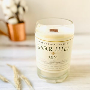 May include: A clear glass candle with a wooden wick. The candle has a cream-colored wax and a label that reads "CALEDONIA SPIRITS BARR HILL GIN" with a bee illustration. The candle is on a white wooden surface.