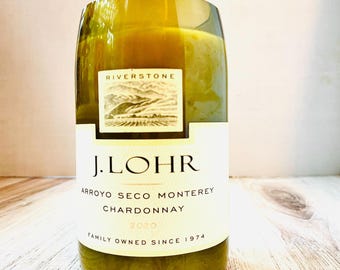 May include: A repurposed wine bottle candle with a white label featuring the text "J. LOHR Chardonnay 2020". The label also includes the text "Riverstone" and "Family Owned Since 1974". The candle has a light green hue.