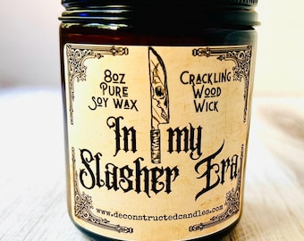 May include: A dark brown glass jar candle with a black lid. The label reads "In my Slasher Era" with a knife graphic. It contains 8 oz of pure soy wax and a crackling wood wick.