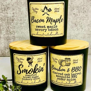 Può includere: Tre barattoli di candele neri con coperchi in legno. Le etichette sono di colore crema con testo nero. Le candele sono etichettate "Bacon Maple", "Smokin'" e "Bourbon & BBQ".