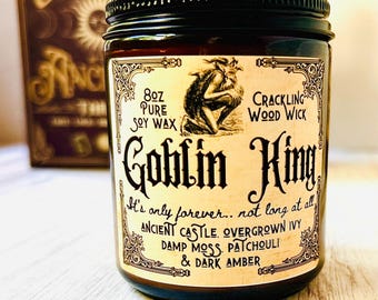May include: A glass jar candle with a black lid and a label featuring the words "Goblin King." The label also includes the text "8oz Pure Soy Wax" and "Crackling Wood Wick." The candle has a dark amber color.