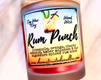 May include: A frosted glass candle with a wooden lid, labeled "Rum Punch." The label features a cocktail illustration and lists ingredients like pineapple, orange, and cherry. The candle is 10 oz and has a wood wick.
