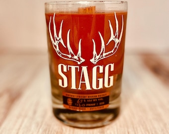 May include: A clear glass candle with a red wax interior. The candle features a white antler design above the word "STAGG" in a bold font. Below the text is a label with additional text. The candle has a wooden wick.