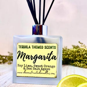 Op de afbeelding: Een heldere glazen fles met een zilveren dop en zwarte diffuserstokjes. De fles heeft een etiket met de tekst "TEQUILA THEMED SCENTS Margarita Key Lime, Sweet Orange & Sea Salt Agave www.deconstructedcandles.com".