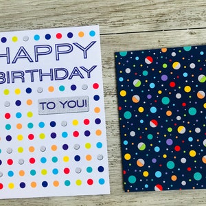 Puede incluir: Una tarjeta blanca con un mensaje azul "Feliz cumplea&ntilde;os" y una pegatina plateada "&iexcl;Para ti!". La tarjeta tiene un fondo blanco con un patr&oacute;n de puntos de colores. La tarjeta est&aacute; al lado de una tarjeta azul con un patr&oacute;n de puntos de colores.