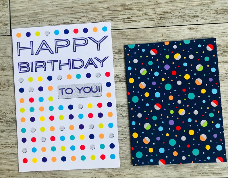 Puede incluir: Una tarjeta blanca con un mensaje azul "Feliz cumplea&ntilde;os" y una pegatina plateada "&iexcl;Para ti!". La tarjeta tiene un fondo blanco con un patr&oacute;n de puntos de colores. La tarjeta est&aacute; al lado de una tarjeta azul con un patr&oacute;n de puntos de colores.