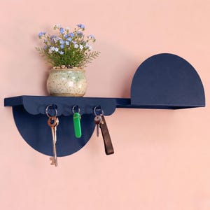 May include: A navy blue metal wall shelf with a semi-circular design, featuring a small shelf and key hooks. A small potted plant with blue flowers sits on the shelf. Three keys hang from the hooks, with a green key and a key with a leather fob.