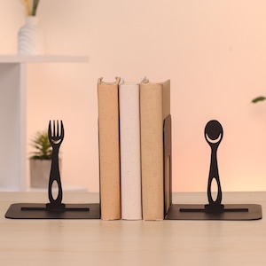 Op de afbeelding: Zwarte metalen boekensteunen met een vork- en lepelontwerp. De boekensteunen houden drie boeken met bruine en witte kaften vast. De vork en lepel hebben een lachend gezichtsontwerp. De boekensteunen staan op een houten oppervlak.