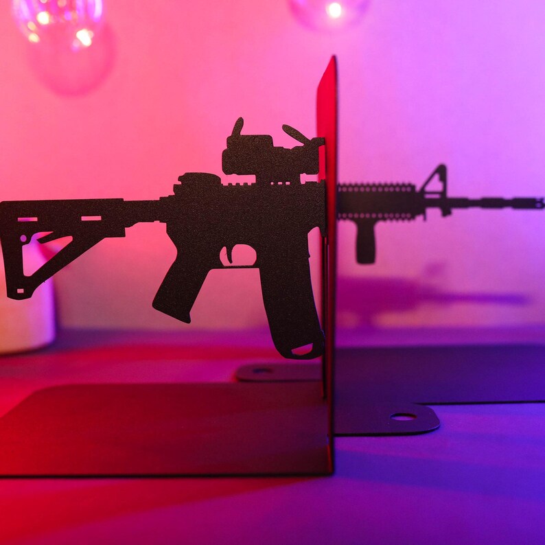 May include: Black metal bookends shaped like a rifle with a scope. The bookends are on a pink and purple surface.