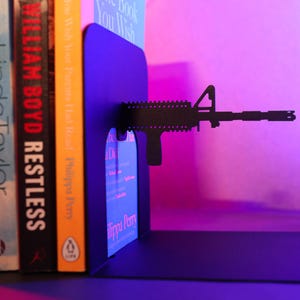 May include: Black metal bookends shaped like a rifle, holding up two books. The bookends are on a shelf with a pink and purple background.