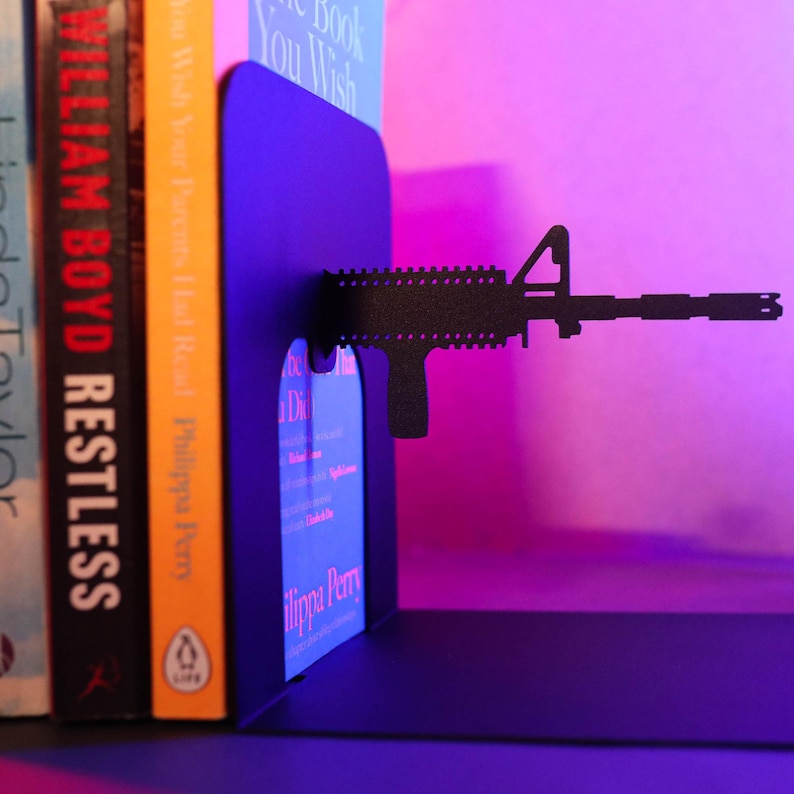 May include: Black metal bookends shaped like a rifle, holding up two books. The bookends are on a shelf with a pink and purple background.