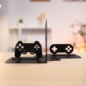 May include: Black metal bookends in the shape of video game controllers. One bookend features a detailed controller design with buttons and directional pad, while the other has a simplified design. The bookends are on a light-colored wooden surface.