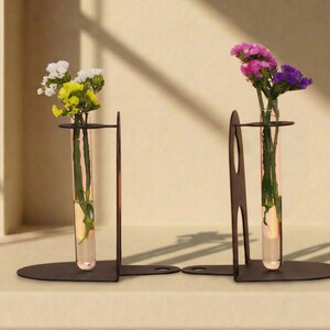 May include: Two black metal bookends, each holding a glass test tube vase with flowers. One vase has white and yellow flowers, the other has purple and pink flowers. The vases are filled with water and sit on a beige surface.