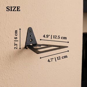 Pu&ograve; includere: Una staffa per mensola da parete in metallo nero con base triangolare e braccio di supporto piatto. La staffa presenta dimensioni di 6 cm di altezza, 12,5 cm di profondit&agrave; e 12 cm di larghezza, come indicato dalle etichette nell'immagine.