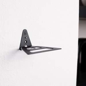 Pu&ograve; includere: Una staffa per mensola a parete in metallo nero opaco, caratterizzata da una piastra di montaggio verticale triangolare con due fori per viti e una piattaforma orizzontale con un design traforato. La staffa &egrave; fissata con una vite.