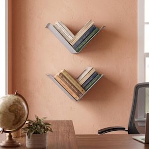 Op de afbeelding: Twee V-vormige wandplanken van lichtgrijs metaal, elk met een verzameling boeken. De boeken hebben verschillende kleuren ruggen met zichtbare titels zoals The Shape of Stories, Kinetic Shelving en The Power of Minimalism.