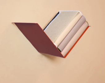 Op de afbeelding: Een V-vormige metalen wandplank met een dieprode buitenkant en een feloranje binnenrand. De plank bevat drie gestapelde rechthoekige voorwerpen met gladde, neutrale oppervlakken in tinten crème, lichtbeige en zacht mauve.