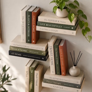以下が含まれることがあります： The Kinfolk Home、This Is Home、Plant Therapyなどのタイトルが記された、ニュートラル、オリーブグリーン、テラコッタカラーの表紙を持つ装飾用書籍のコレクション。壁掛け棚に配置され、鉢植えとリードディフューザーが添えられています。