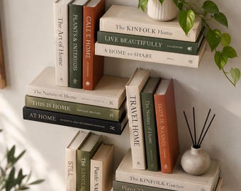 Op de afbeelding: Een verzameling decoratieve boeken op wandplanken, met neutrale, olijfgroene en terracotta covers met titels zoals The Kinfolk Home, This Is Home en Plant Therapy, vergezeld door een kamerplant en een geurstokjeshouder.