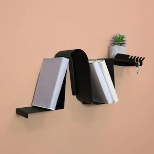May include: A black metal wall shelf with a unique, wavy design. The shelf holds several books with white pages and a small potted plant. A section of the shelf has hooks for keys. The shelf is mounted on a peach-colored wall.