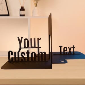 May include: Black metal bookends with the words "Your Custom" and "Text" cut out. The bookends are on a light-colored wooden surface. The design is modern and minimalist, suitable for home or office decor.