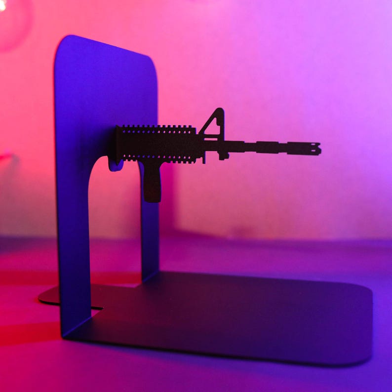 May include: A black metal bookend shaped like a rifle. The bookend is on a pink and purple surface.