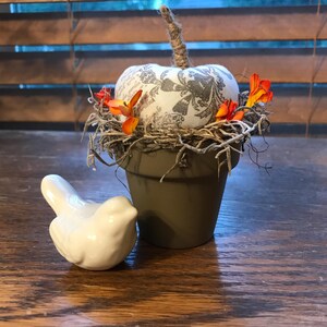 May include: A small gray and white fabric pumpkin sits in a brown terracotta pot. The pot is surrounded by brown twigs and orange flowers. A white ceramic bird sits next to the pot.