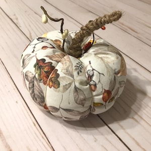May include: A fabric pumpkin decoration featuring a cream-colored base with a printed pattern of brown, orange, and green autumn leaves and acorns. The pumpkin has a twisted jute rope stem and thin wire accents with small white berries.