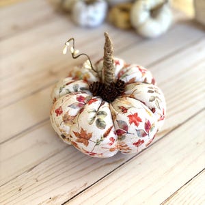 May include: A fabric pumpkin featuring a cream-coloured base with a printed pattern of colourful autumn leaves, berries, and foliage. The pumpkin has a central stem wrapped in twine, accented with small brown pinecones and a thin wire twig.