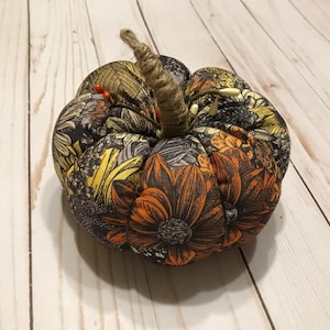 May include: A small, fabric pumpkin with a floral pattern in shades of orange, yellow, and brown. The pumpkin has a brown stem and is sitting on a white wooden surface.