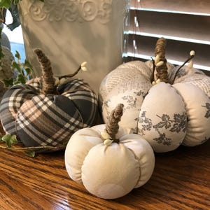 May include: Three decorative fabric pumpkins in various patterns. The pumpkins are beige, gray, and white with plaid and floral patterns. The pumpkins have brown stems with white accents.