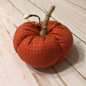 May include: A small orange pumpkin with gold polka dots and a hessian stem. The pumpkin is sitting on a light wood surface.