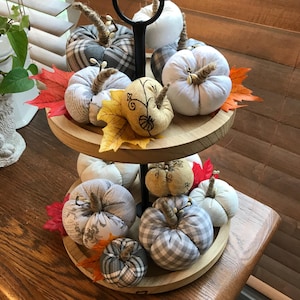 May include: A two-tiered wooden stand with a black metal base holds a collection of fabric pumpkins in various patterns and colors, including plaid, floral, and solid white. The pumpkins are decorated with fall leaves and have a rustic, farmhouse aesthetic.